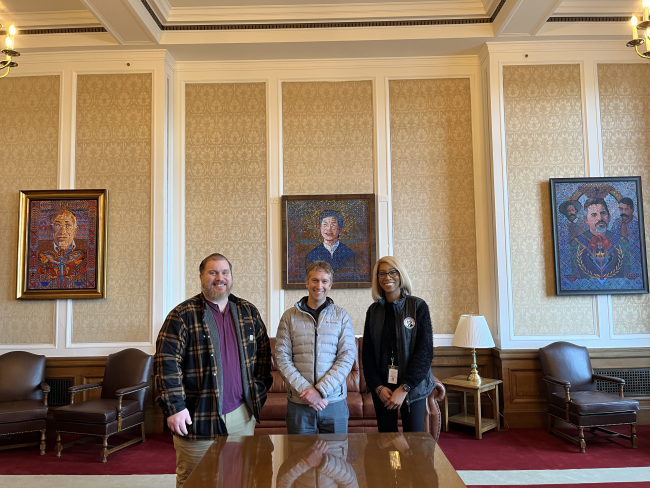 Three people smiling for a picture in front of three oil painting portraits inside a room with a high ceiling, white and beige walls, a dark red carpet, and old fashioned furniture.