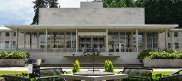 A view of the Joel Pritchard State Library's front side, which is mainly made of white stone with large glass walls. A fountain, a sundial, four metal park benches, and various green bushes are in front of the building.