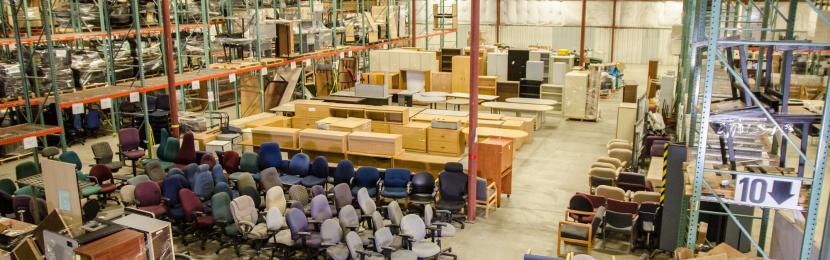 A high-angle view of various office equipment, including rolling chairs, cabinets, and desks, organized on a well-lit warehouse floor.