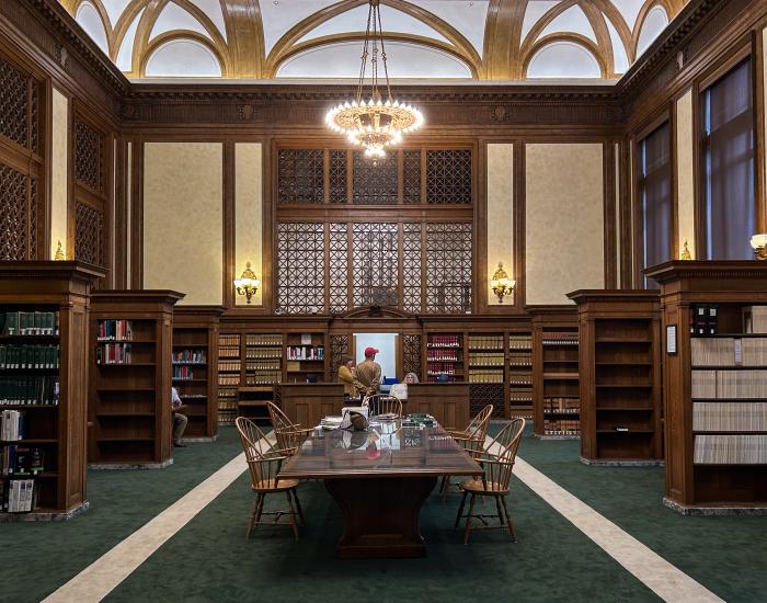 The Washington Law Library lobby, which has a dark green carpet and a brown wooden table in the middle of the room, with brown wooden bookshelves on both sides of the table. A large chandelier hangs above the front desk, where a person is talking to two clerks. 