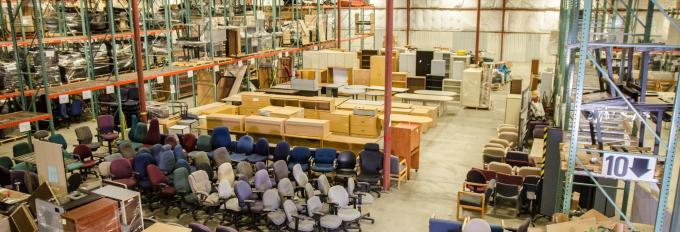 A high-angle view of various office equipment, including rolling chairs, cabinets, and desks, organized on a well-lit warehouse floor.